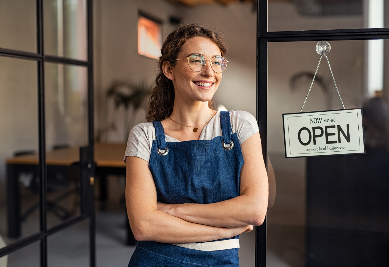 A female small business owner standing with a smile and her arms crossed in front of her business with a "Now Open" sign behind her.